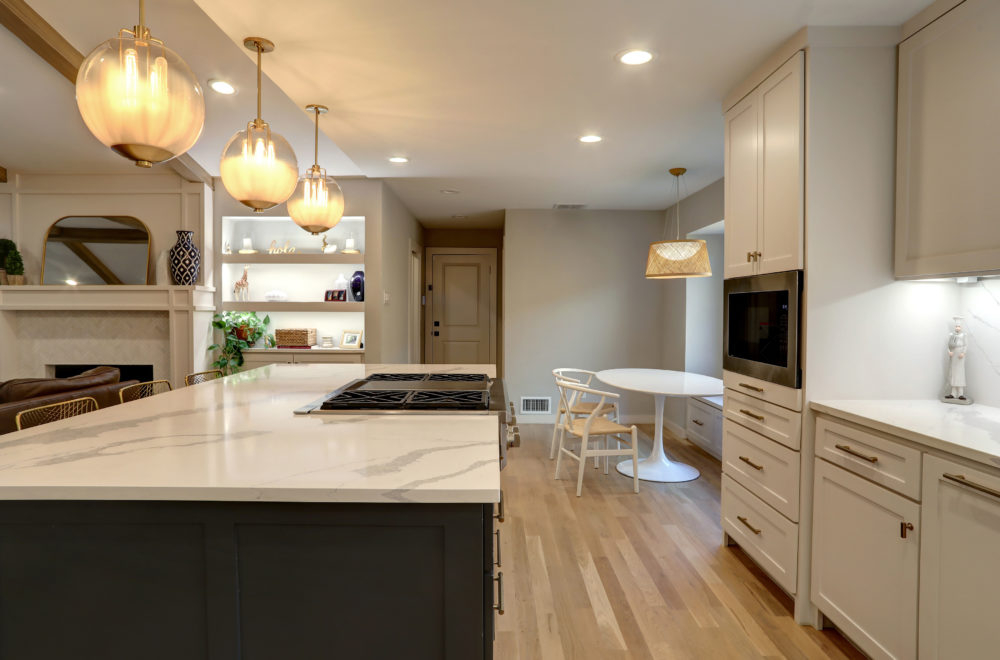 Modern kitchen with marble countertops, pendant lighting, light wood floors, white cabinets, a breakfast nook with a round table, and a partial view of a living area with built-in shelves.