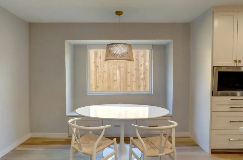 A minimalist dining area with a round white table, two white chairs, a woven pendant light, and a window with a wooden fence view.