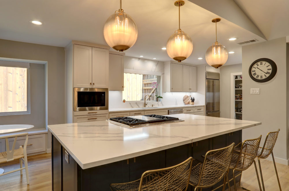 Modern kitchen with a large marble island, built-in appliances, three pendant lights, gold wire barstools, and a wall clock. Natural light comes through windows.