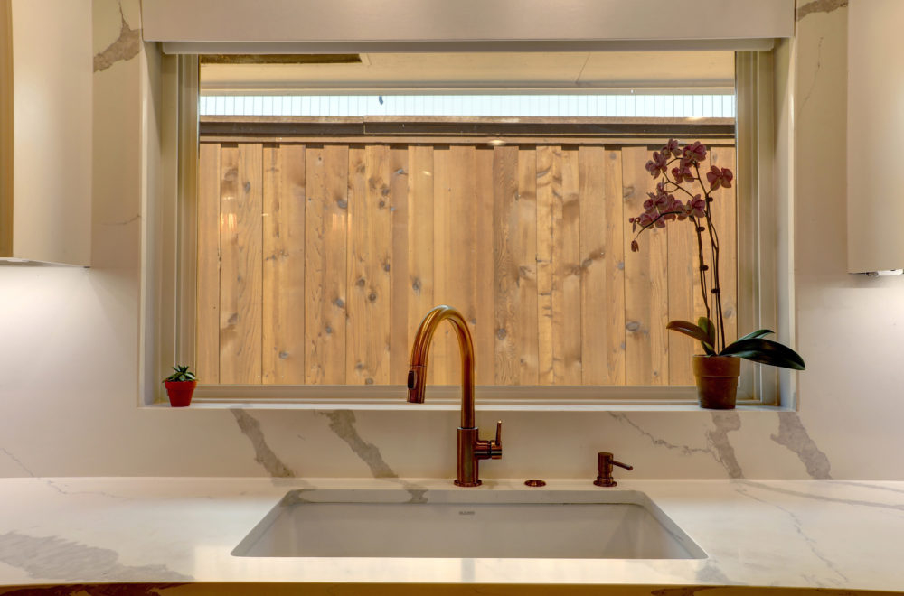 Millridge-16-1000x660 A kitchen sink with a copper faucet is set in a marble countertop beneath a window with a view of a wooden fence; two potted plants sit on the windowsill.