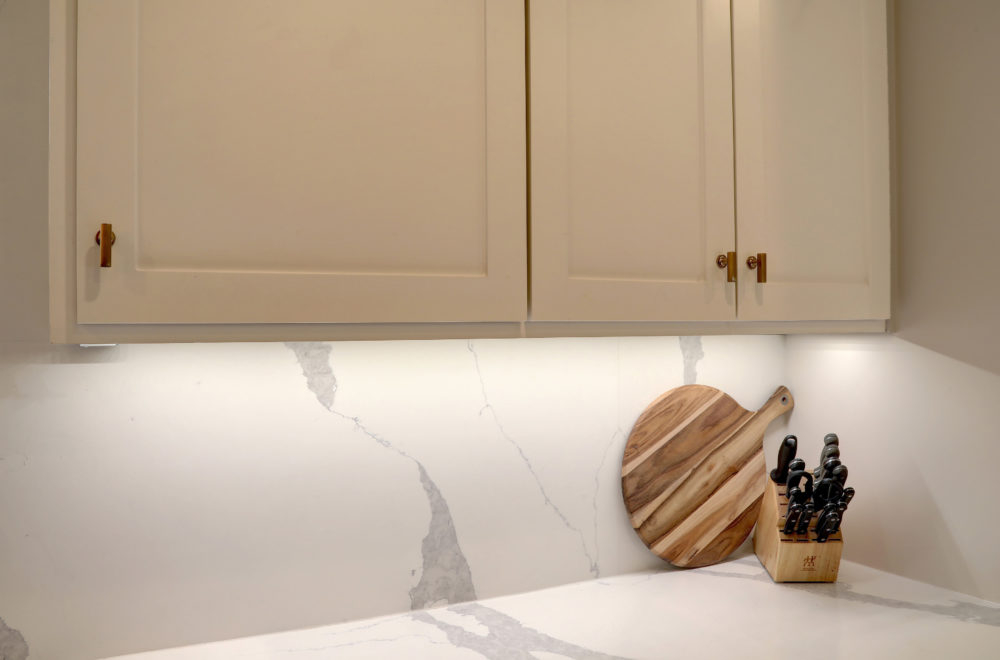 A kitchen counter with white cabinets above, a round wooden cutting board, and a block holding kitchen knives against a white marble backsplash.