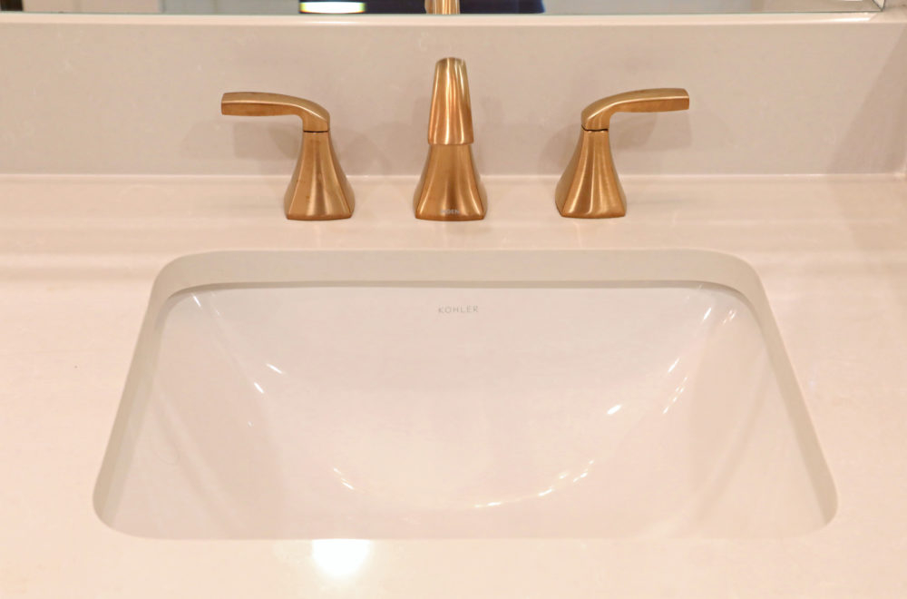 White bathroom sink with a rectangular basin and three brushed gold fixtures for hot, cold, and faucet, set in a light countertop.