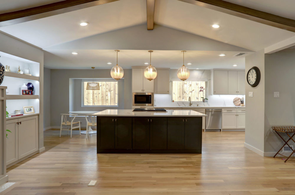 Modern kitchen with light wood floors, a central island with dark cabinets, pendant lights, white cabinetry, and open shelving. Dining area and window are visible in the background.