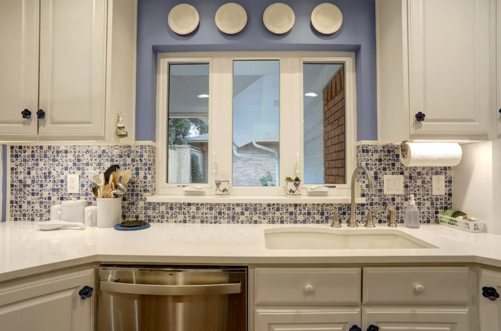 A modern kitchen remodel Dallas with white cabinets, blue mosaic tile backsplash, stainless steel dishwasher, and a window above the sink.