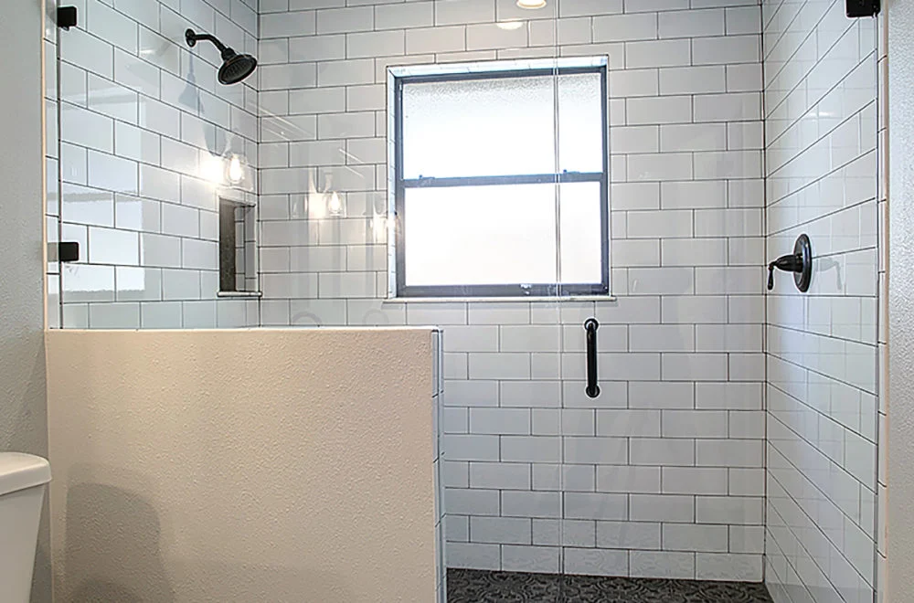 A walk-in shower with white subway tiles, a glass divider, a window, two shower heads, and a black grab bar on the wall.