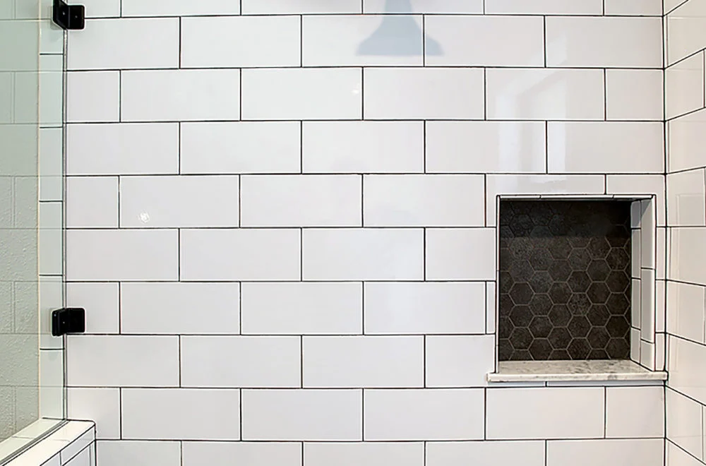 White subway tile shower wall with black grout lines, a glass door with black hardware, and a recessed shelf lined with dark hexagonal tiles and a marble shelf.