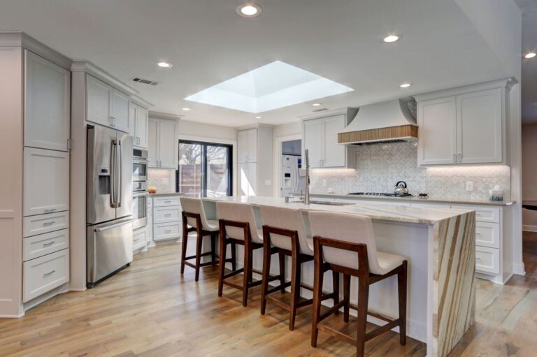 Modern kitchen with white cabinets, stainless steel appliances, a large island with a marble waterfall countertop, five bar stools, wood flooring, and a skylight above.