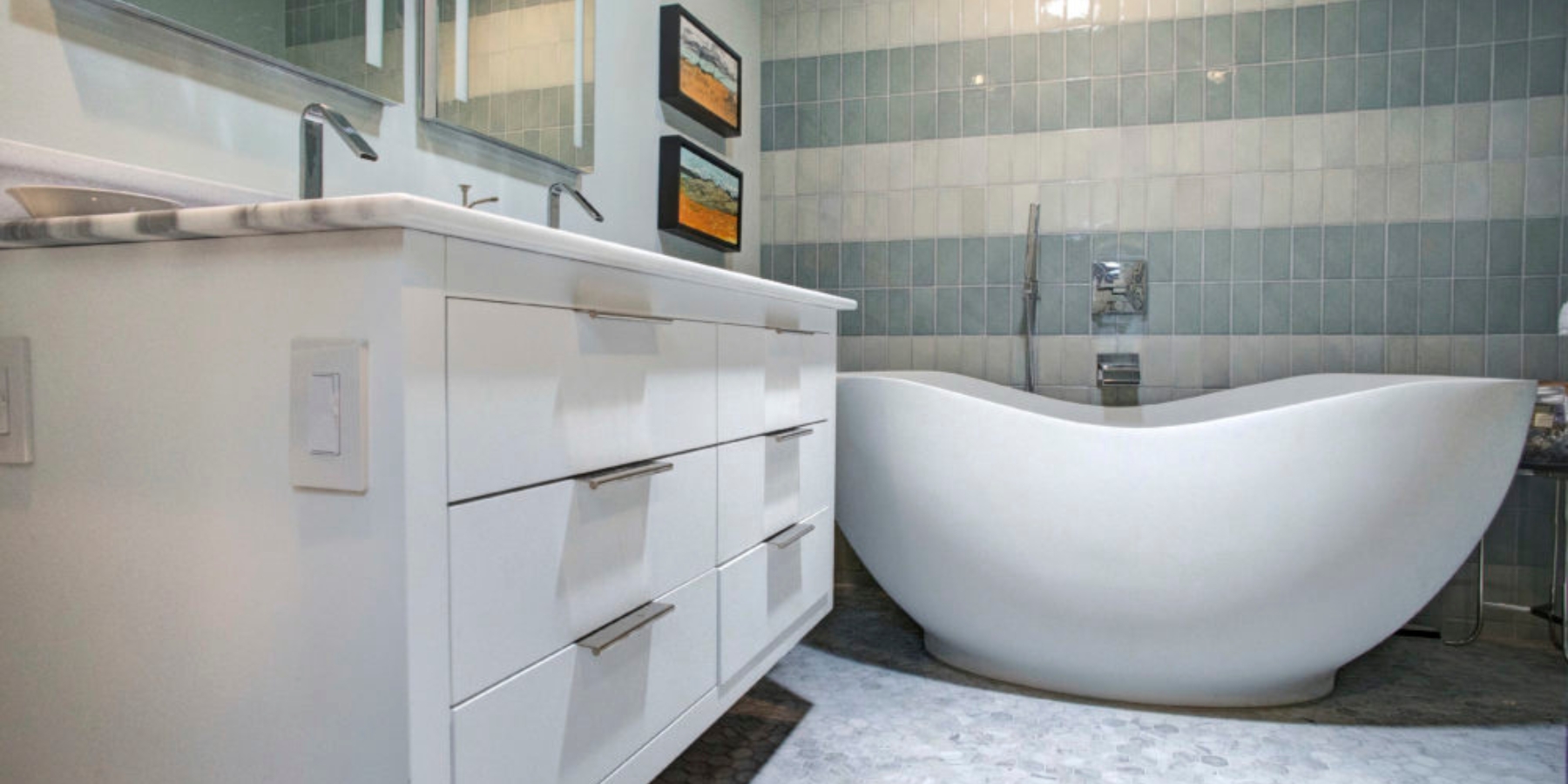 Modern bathroom with a white freestanding bathtub, a white vanity with drawers, a large mirror, and two small wall art pieces above the tub.
