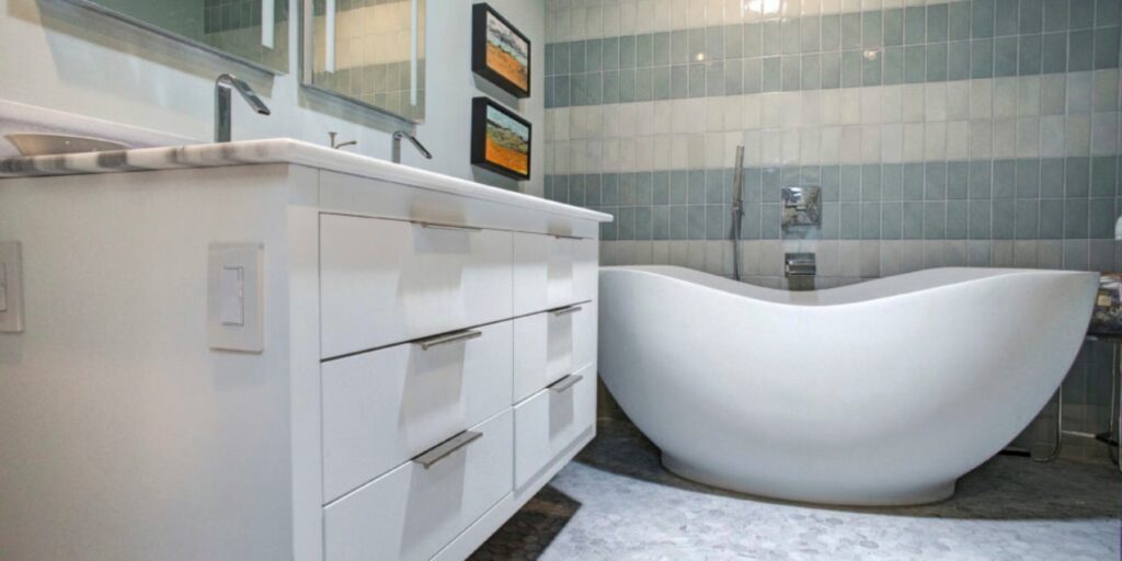 Modern bathroom with a white freestanding bathtub, white vanity with drawers, dual sinks, wall tiles in vertical stripes, and two small framed artworks above the tub.