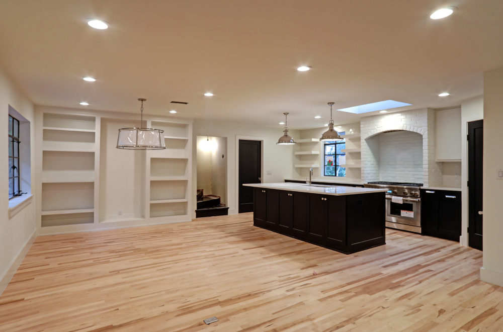 Spacious open-plan kitchen and living area with light wood floors, built-in shelves, large island, modern lighting, and white walls.