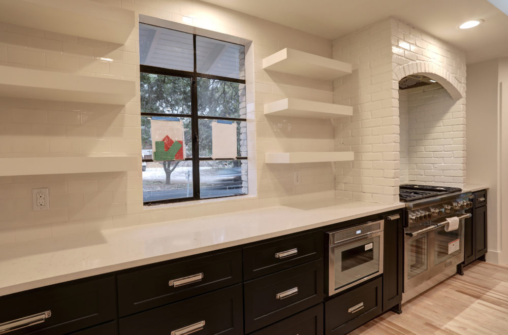 Modern kitchen with black lower cabinets, white countertops, open white shelves, a stainless steel oven and microwave, and a large window above the counter.
