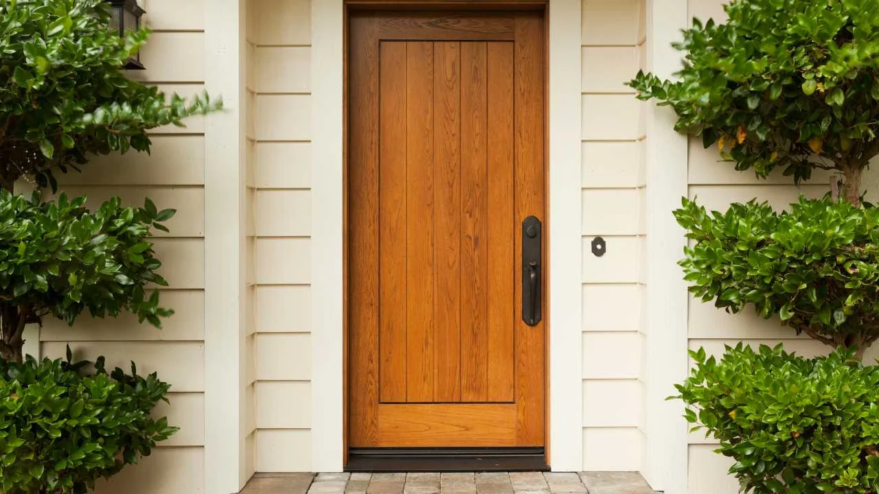 Wooded Door to House A wooden front door with a vertical panel design, surrounded by cream-colored siding and flanked by neatly trimmed green bushes.
