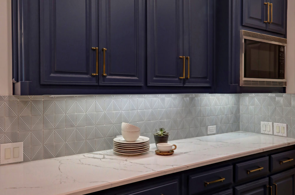 A modern kitchen countertop with blue cabinets, gold handles, a marble-patterned surface, stacked dishes, a small plant, and a built-in microwave.