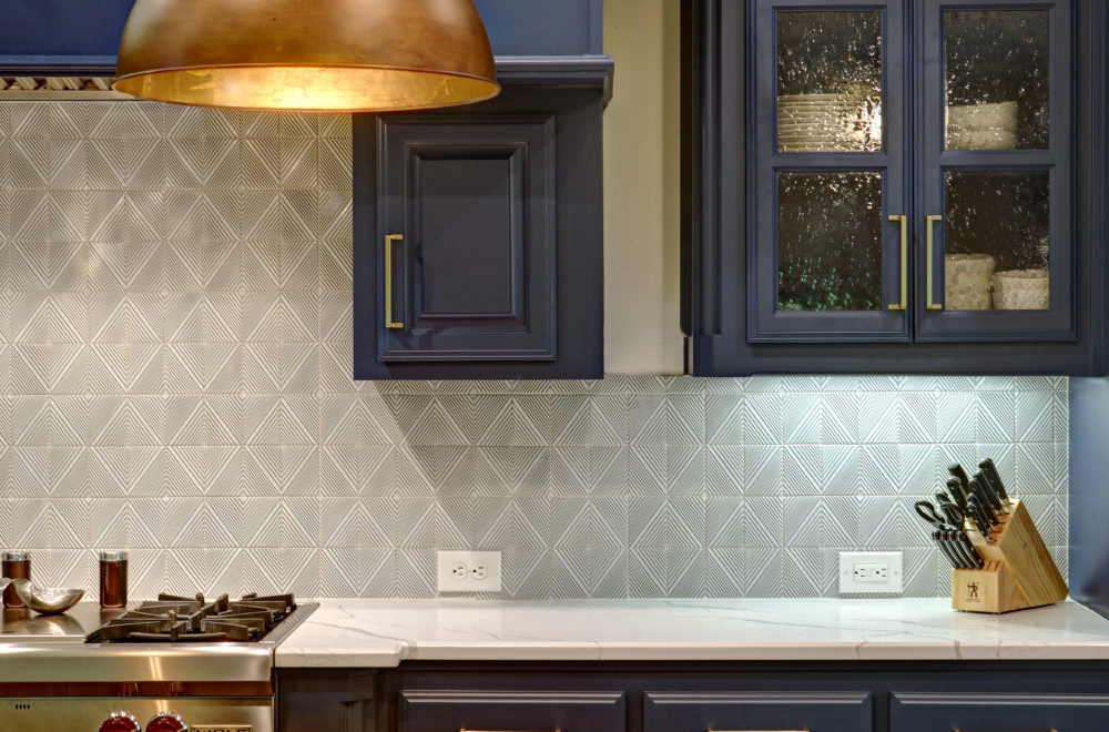 A modern kitchen with navy cabinets, a patterned tile backsplash, marble countertops, a stove, a knife block, and under-cabinet lighting.