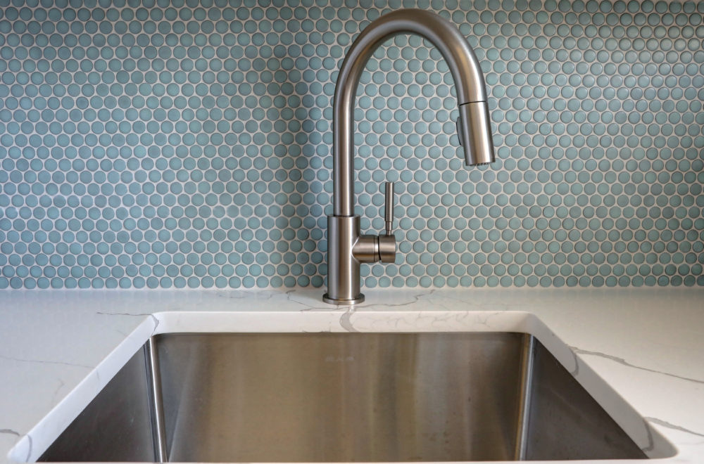A stainless steel kitchen sink with a modern curved faucet, set against a blue hexagonal tile backsplash and white countertop.
