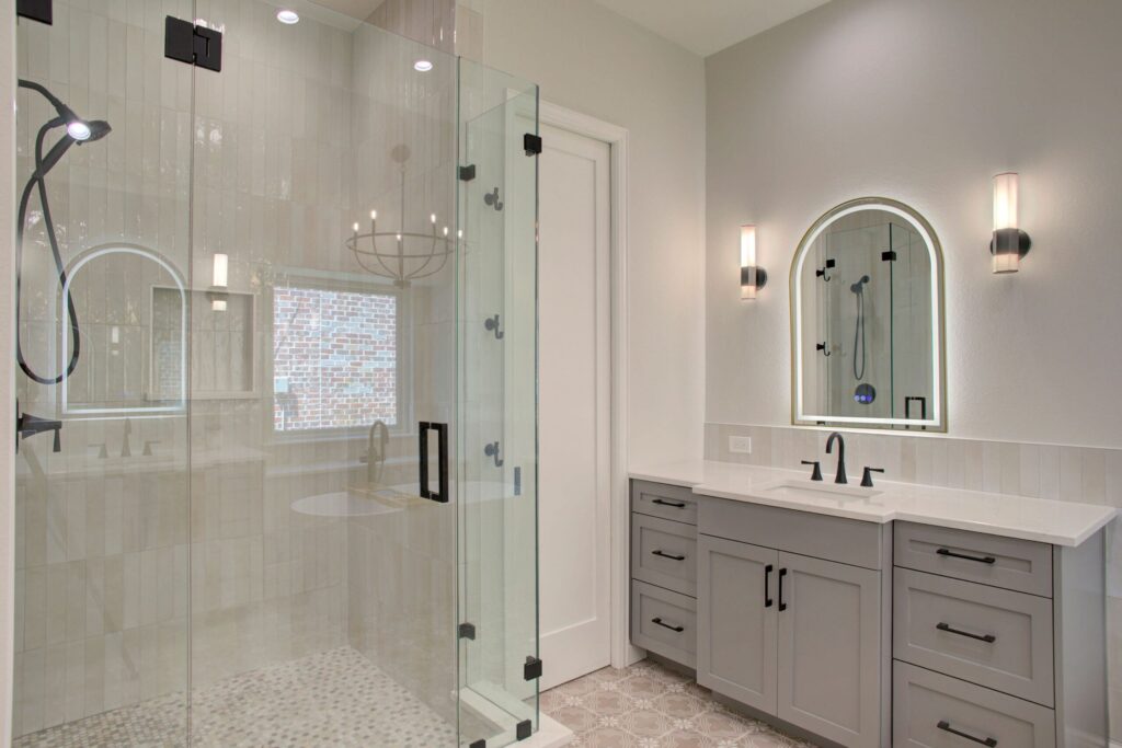 Modern bathroom with a glass-enclosed shower, a double-sink vanity with gray cabinets, wall sconces, and an arched mirror.