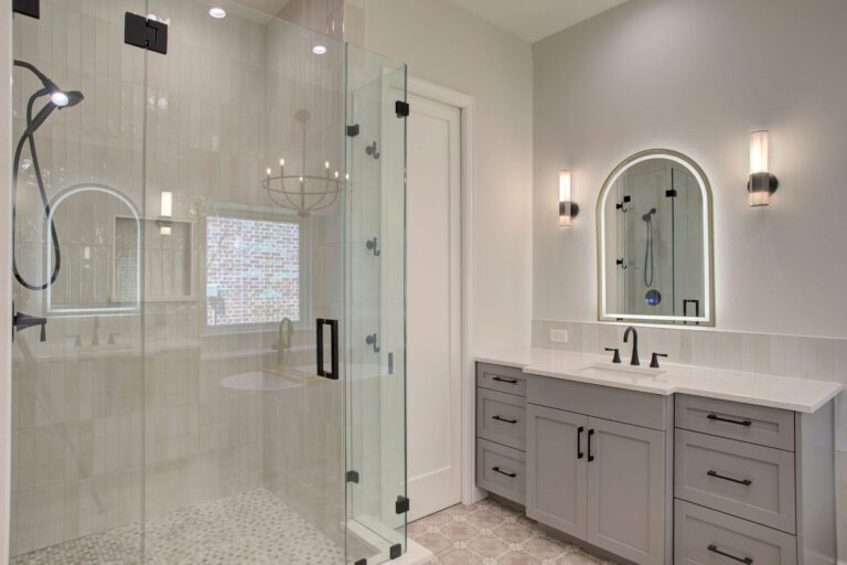 Modern bathroom with a glass-enclosed shower, a double-sink vanity with gray cabinets, wall sconces, and an arched mirror.