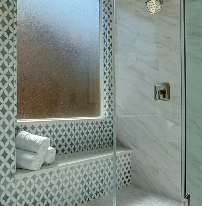 A shower with a built-in tiled bench, patterned blue and white wall tiles, a glass door, and three rolled white towels on the bench next to a frosted window.