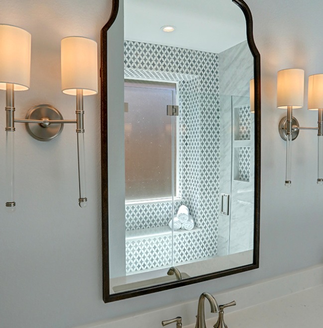 A bathroom mirror reflects a glass shower with patterned tile. Two wall-mounted light fixtures with white shades are visible on either side of the mirror.