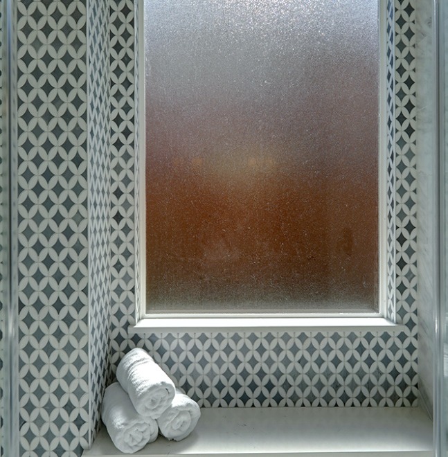 A frosted window set in a patterned tile wall with three rolled white towels stacked in the corner of the windowsill.