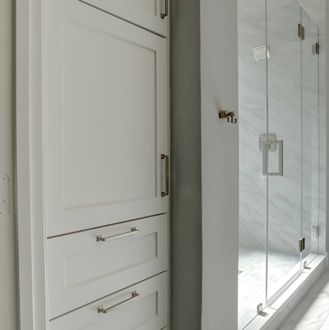 White built-in cabinetry with silver handles next to a glass-enclosed shower with marble walls in a bathroom.