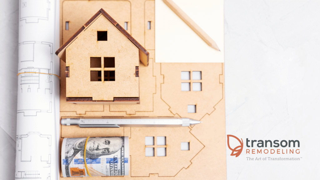 A model house, blueprint, pencil, pen, and rolled dollar bill are arranged on a table next to the Transom Remodeling logo.