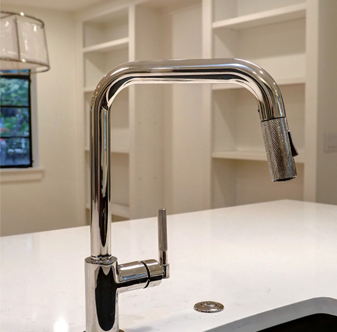 A modern chrome kitchen faucet is installed on a white countertop with empty open shelves and a window visible in the background.