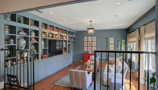 A living room with blue built-in shelves, decorative vases, books, a patterned armchair, a gray sofa, hardwood floors, and large windows with bamboo shades.