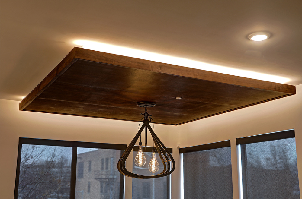 A modern ceiling with a wooden drop panel, recessed lighting, and a decorative light fixture with exposed bulbs in a room with large windows.