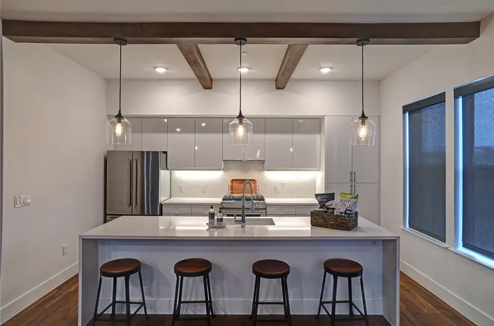 Modern kitchen with a white island, three wooden stools, pendant lights, stainless steel appliances, wood ceiling beams, and large windows with dark shades.