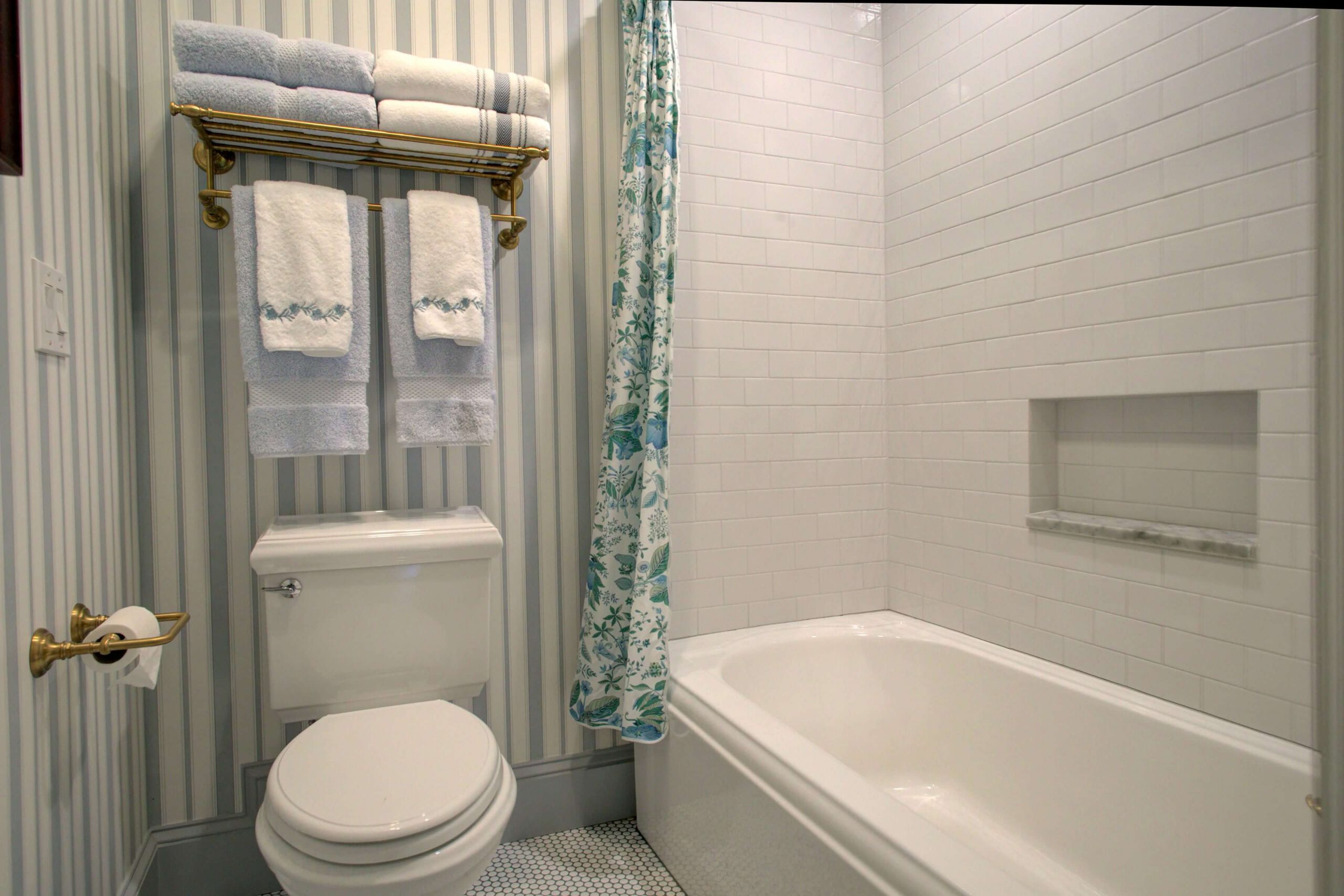 A bathroom with a toilet, bathtub, white tiled walls, a floral shower curtain, and shelves holding folded towels above the toilet.