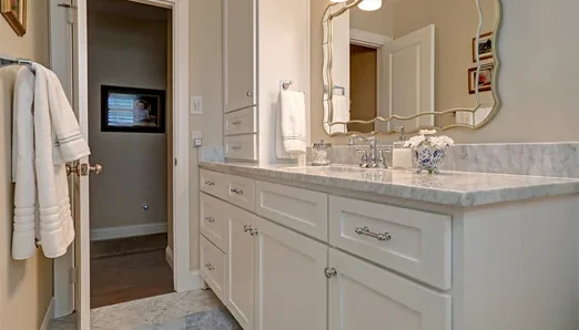 new1-1000x660 Bathroom with white cabinets, marble countertop, gold-framed mirror, and a towel hanging on the wall; doorway opens into a hallway.