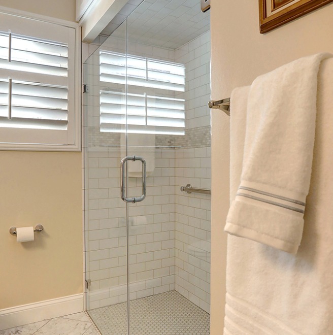 Modern bathroom with glass-enclosed shower, white subway tiles, grab bar, window shutters, towel rack with folded towels, and a toilet paper holder.