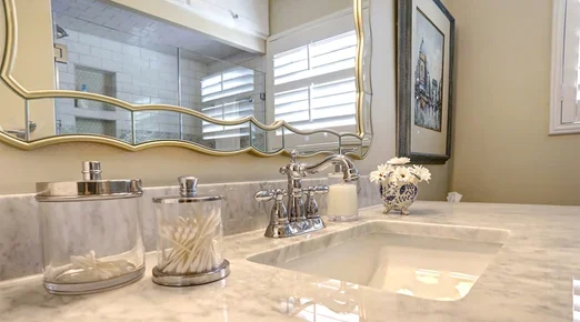 A bathroom vanity with a marble countertop, glass jars holding cotton swabs and pads, decorative flowers, a silver faucet, and a large framed mirror.
