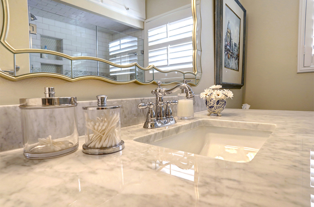 A marble bathroom countertop with a sink, chrome faucet, two glass containers with cotton swabs, a soap dispenser, and a small vase with white flowers.