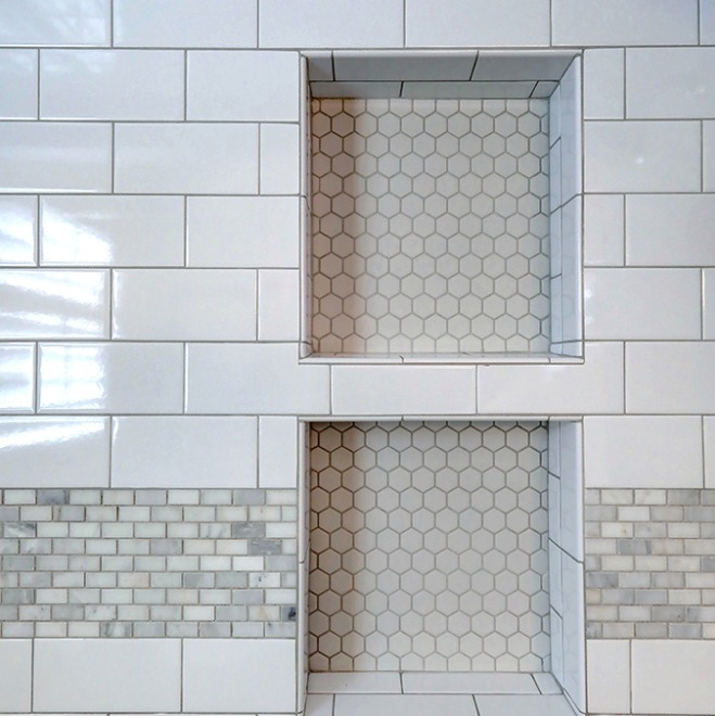 White tiled wall with two recessed shelves featuring small hexagonal tiles, bordered by rectangular subway tiles and a strip of mosaic tiles below each shelf.