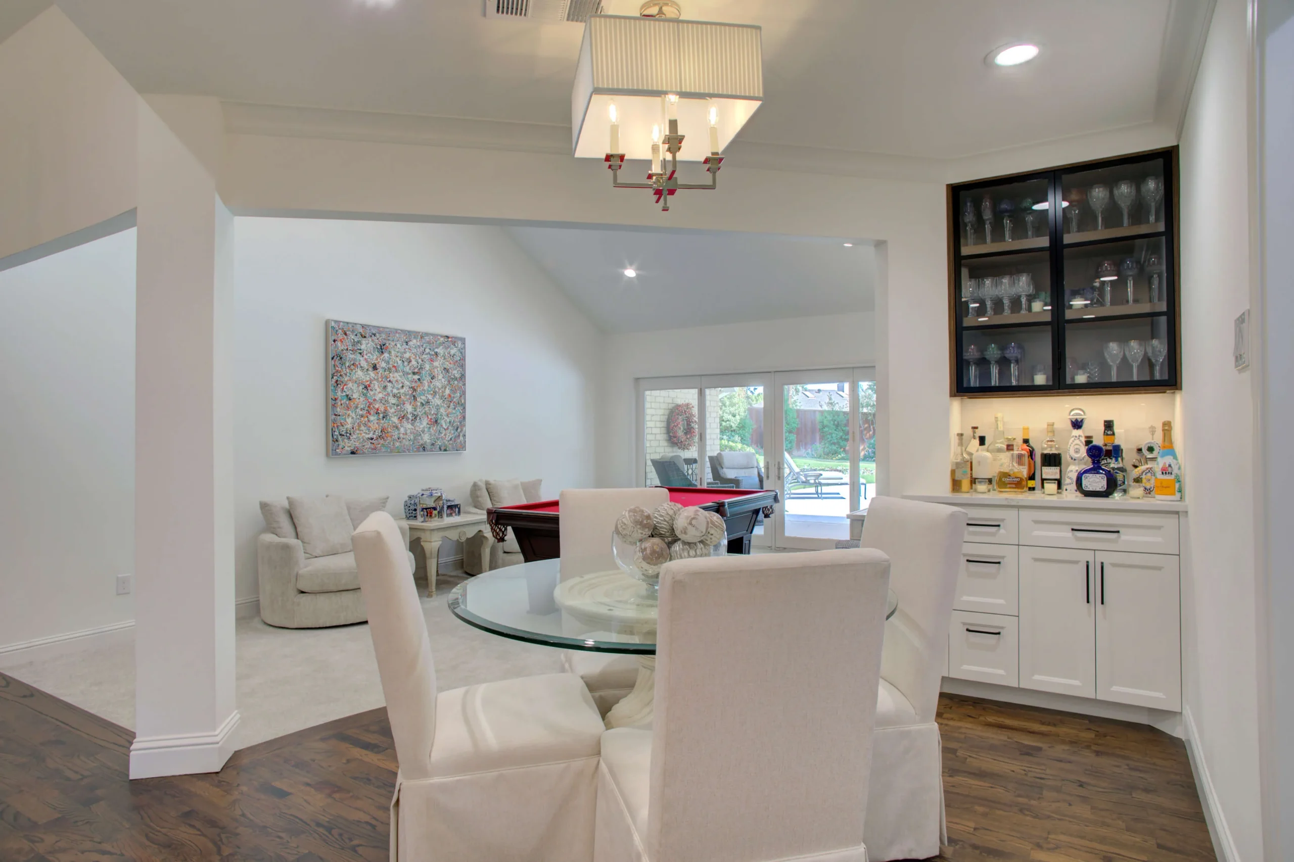 A dining area in a Dallas area home with a round glass table and four white chairs is next to a built-in bar and open living space with a pool table and seating area in a modern home.
