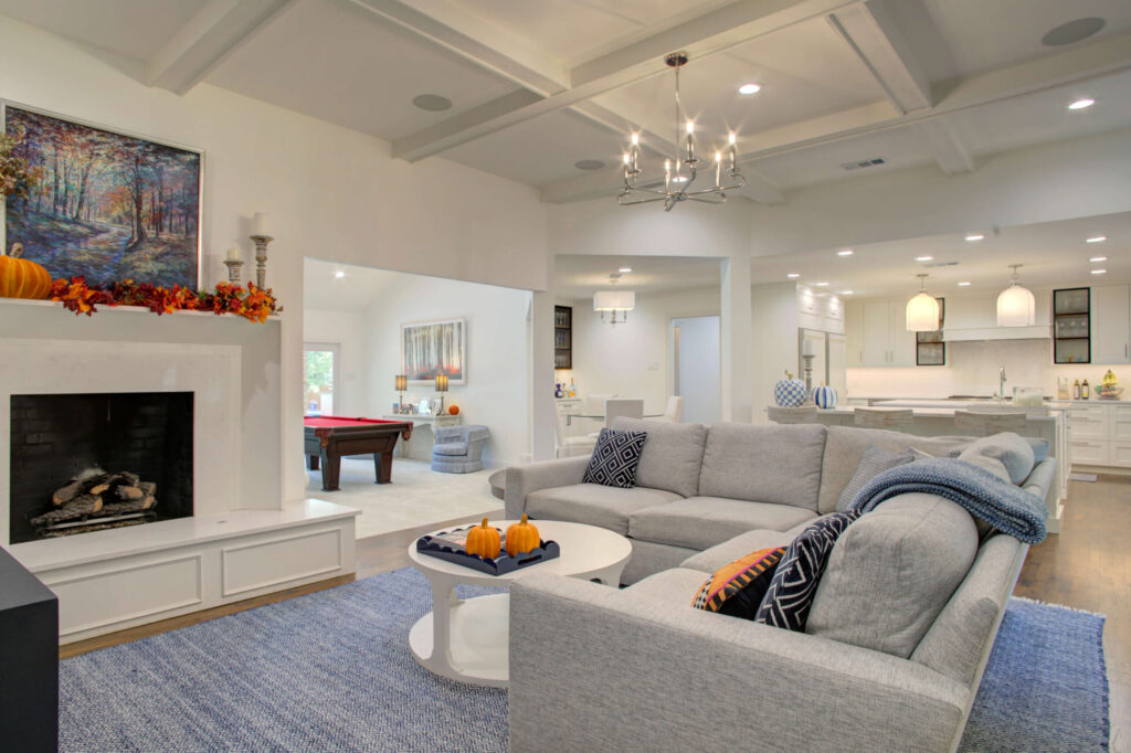 A modern living room with a gray sectional sofa, blue rug, pumpkins on the table, a fireplace decorated with autumn garland, and an open view to a kitchen and game area.