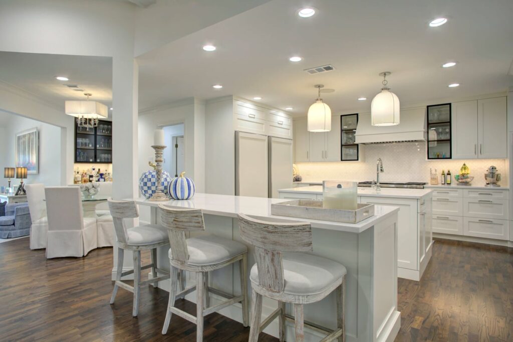 Modern white kitchen with island, three barstools, pendant lights, built-in cabinets, and stainless steel appliances; dining area visible in the background.