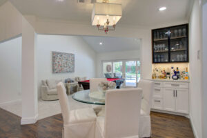 Open dining area with a round glass table and four white chairs, adjacent to a built-in bar and a living space with large windows and a pool table.