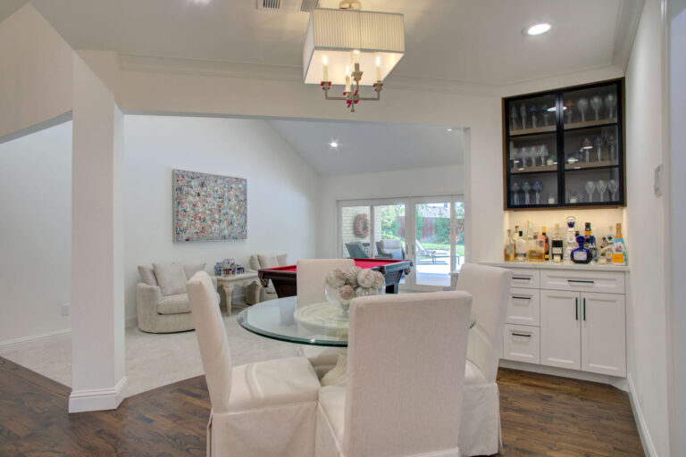 Open dining area with a round glass table and four white chairs, adjacent to a built-in bar and a living space with large windows and a pool table.