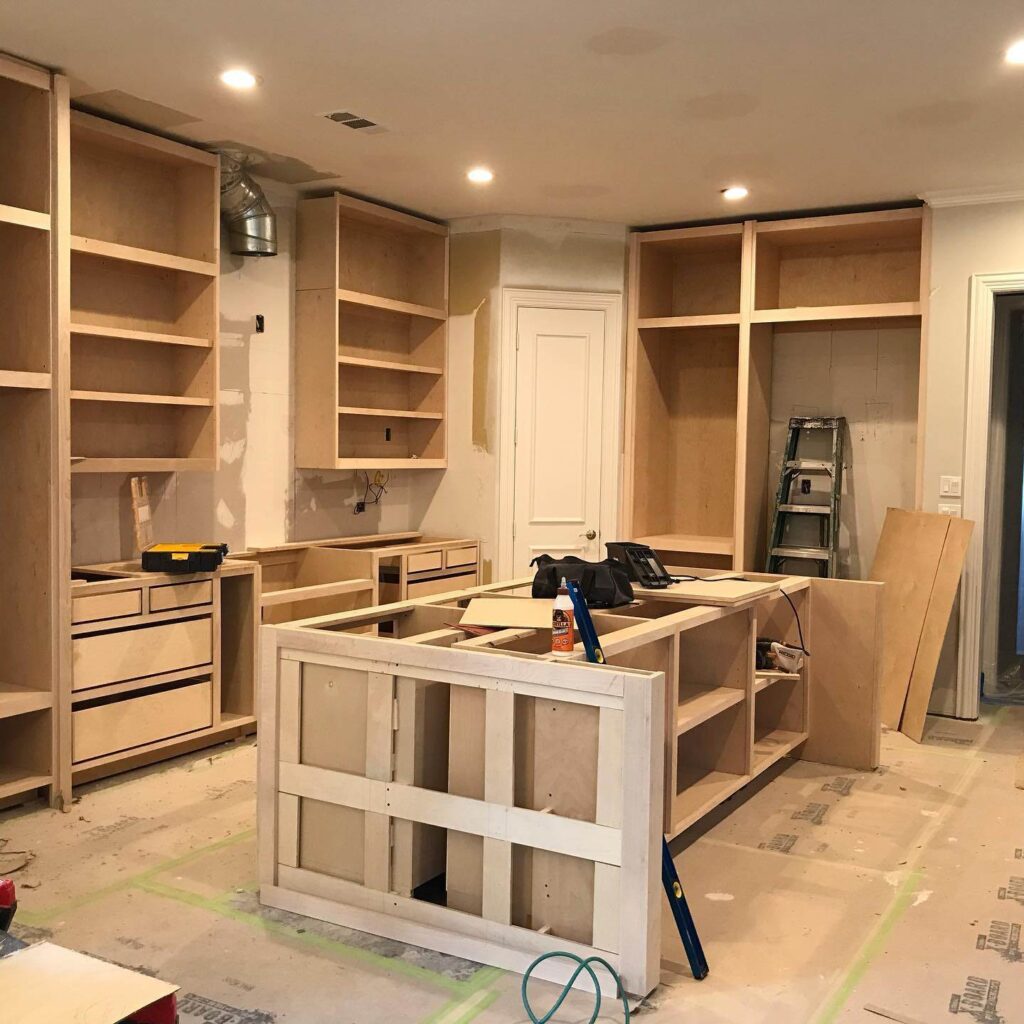 Kitchen under construction with unfinished wooden cabinets, an island, tools, a ladder, and protective floor coverings visible.