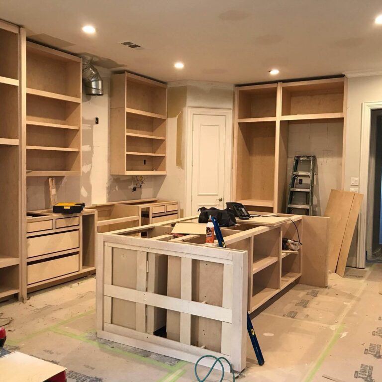 Kitchen under construction with unfinished wooden cabinets, an island, tools, a ladder, and protective floor coverings visible.