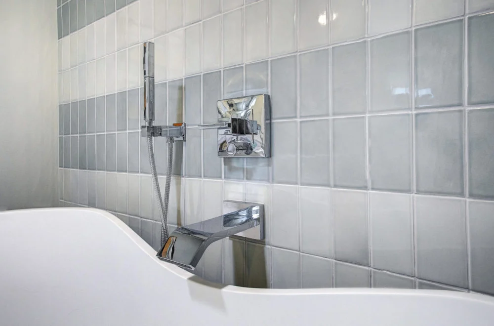 Close-up of a modern bathtub faucet and handheld showerhead mounted on a wall with light gray and white square tiles.