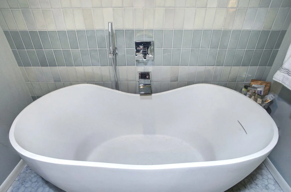 Freestanding white bathtub with modern chrome fixtures against a tiled wall; toiletries are placed on a small table to the right.
