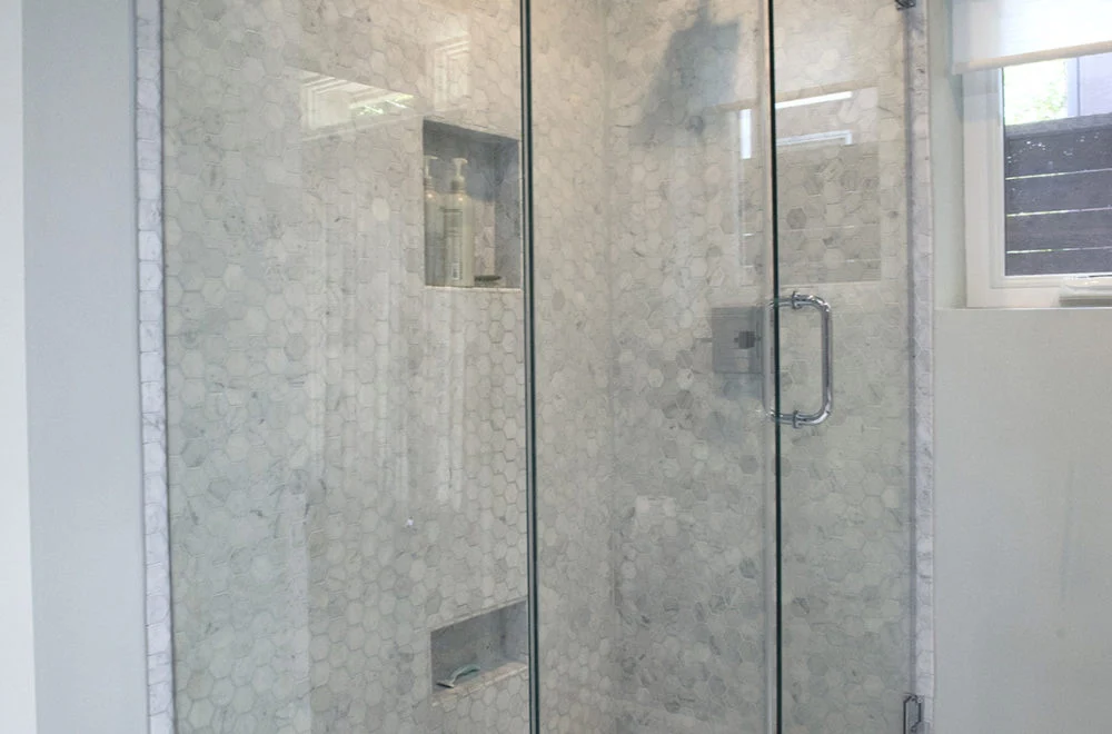Glass shower enclosure with a chrome handle, hexagonal wall tiles, built-in shelves holding bottles, and a window to the right.