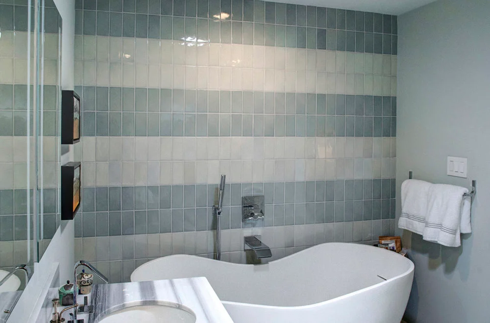 Modern bathroom with gray and white striped tile wall, white freestanding bathtub, towel rack with white towels, and part of a vanity with sink visible.