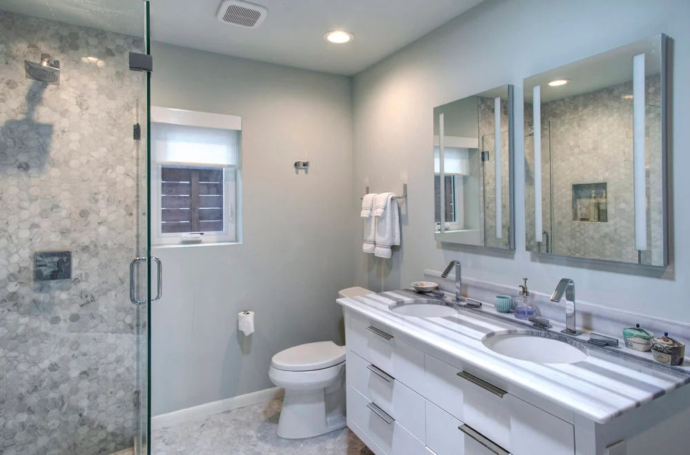 Modern bathroom with a glass-enclosed shower, double-sink vanity, wall mirrors, toilet, and a small window with a shade.