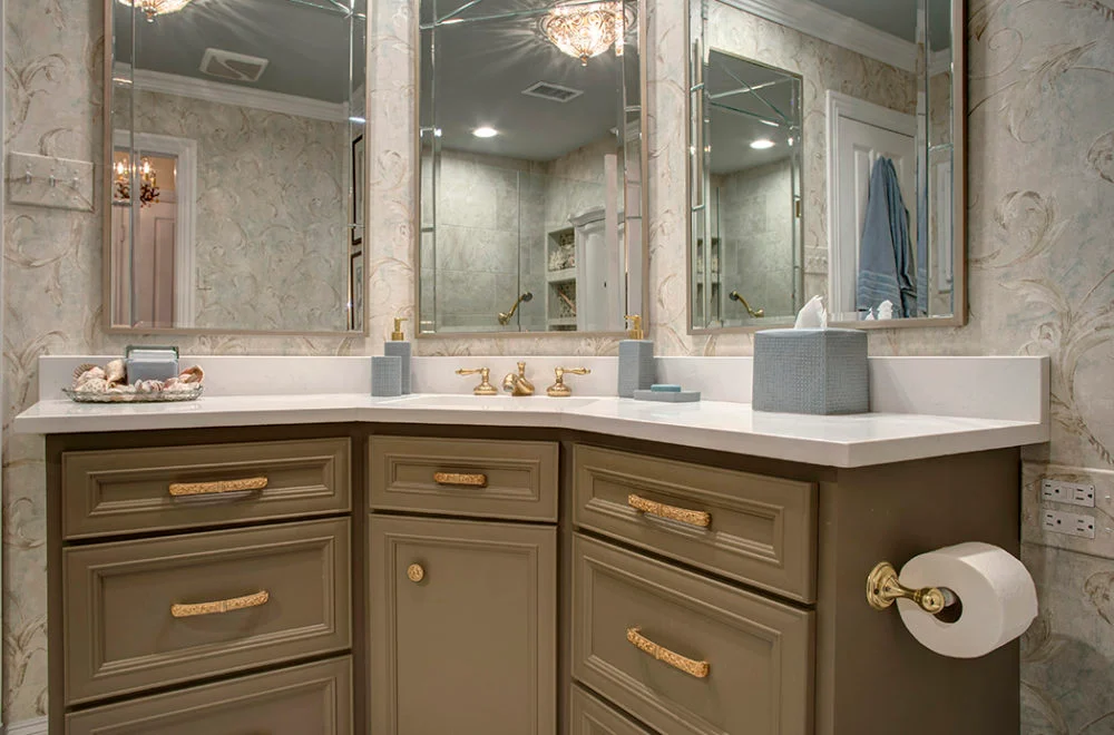Bathroom vanity with gold hardware, dual sinks, wall-mounted mirrors, beige and white wallpaper, and countertop accessories including soap dispensers, a tissue box, and a tray with seashells.