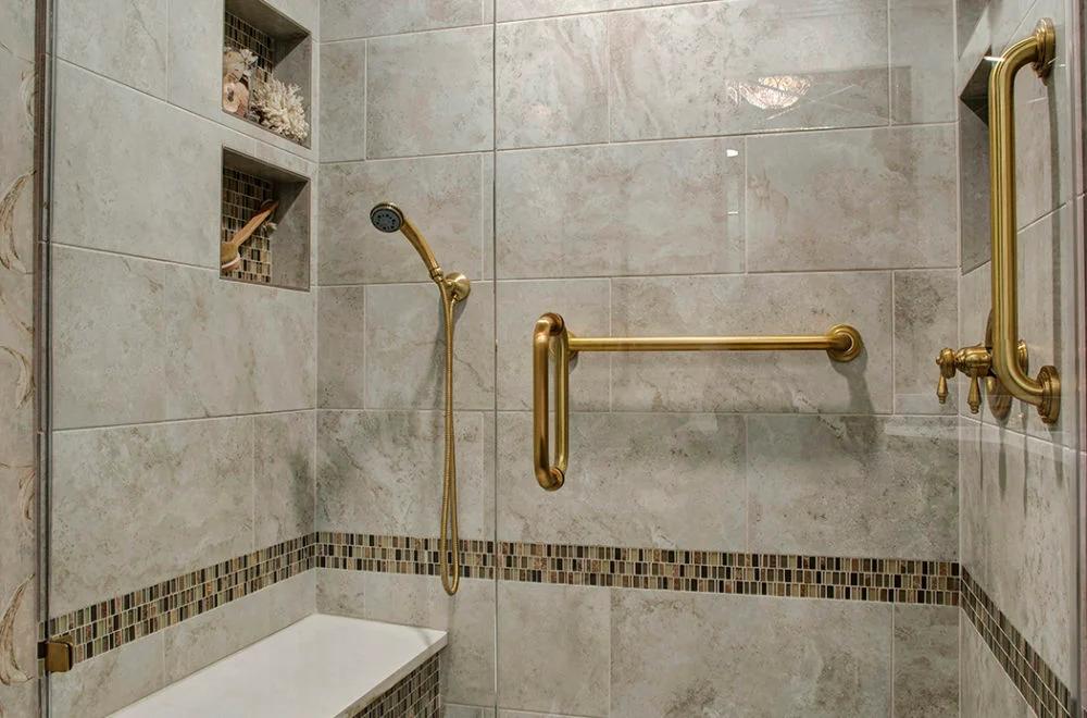 A tiled walk-in shower with a gold handheld showerhead, two gold grab bars, built-in shelves, and a decorative tile border.
